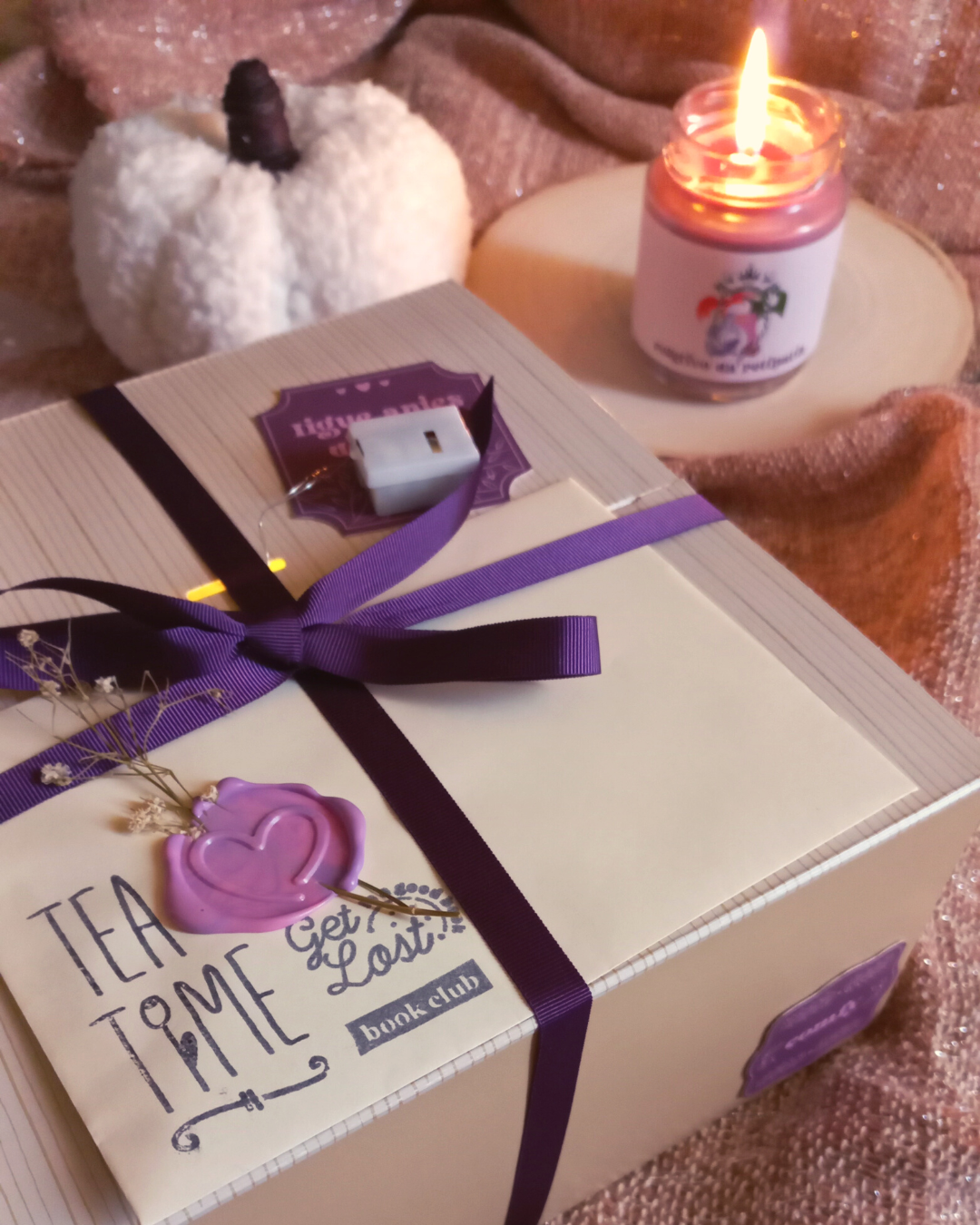 Fotografia de uma caixa literária decorada com uma fita roxa e com um envelope na tampa, ao fundo, uma vela acesa e uma mini abóbora de almofada.