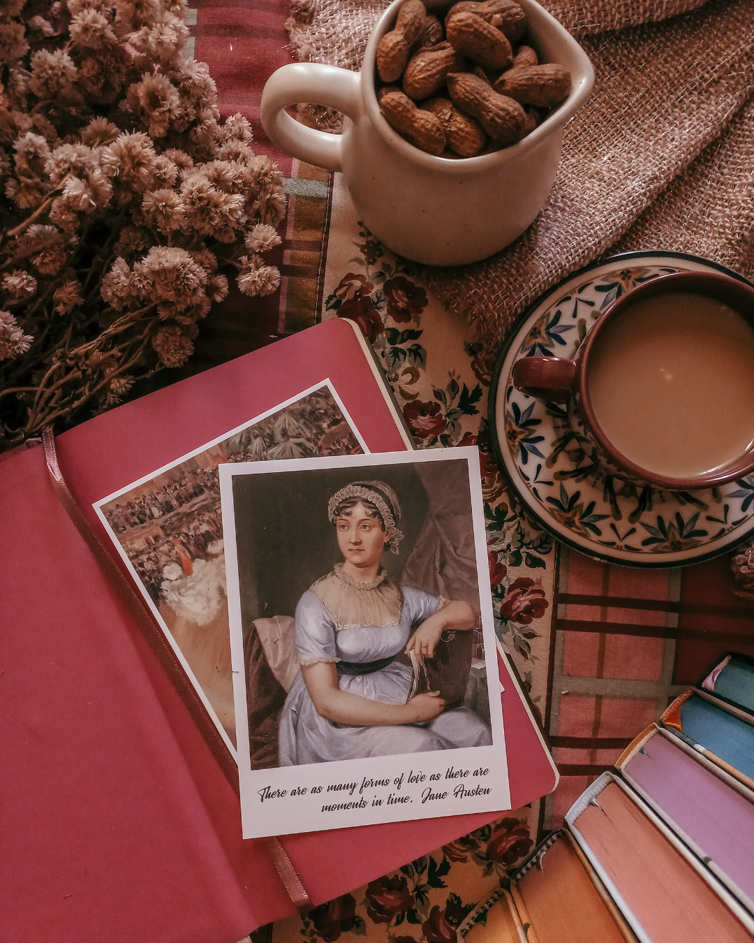 Exemplificando o guia com todos os livros de Jane Austen, a fotografia mostra um cenário para livros em flat lay, com card com fotografia da autora em destaque, sobre um livro aberto e, ao redor, uma pilha de livros mostrando o corte colorido das folhas, uma xícara com café com leite, um jarrinho com amendoim na casca e um ramalhete de flores secas.