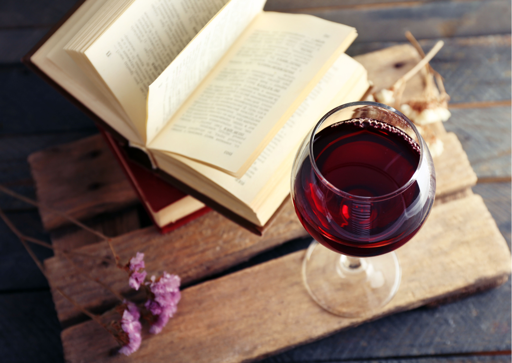 A fotografia em flat lay mostra na imagem um livro aberto e uma taça com vinho, sobre uma tábua de madeira, exemplificando o texto principal da newsletter sobre vinho e literatura.